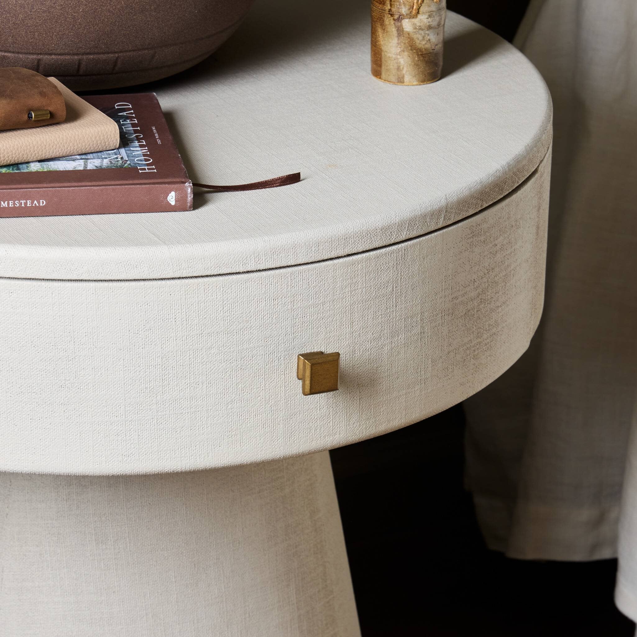 Round beige side table with a gold knob, books, and a candle on top.
