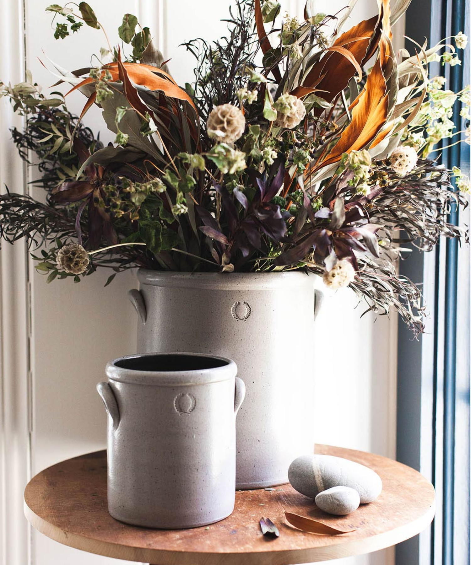 Two gray stoneware crocks of varying sizes with small handles styled with florals. Each crock has a subtle embossed wreath design, conveying a rustic, artisanal feel.