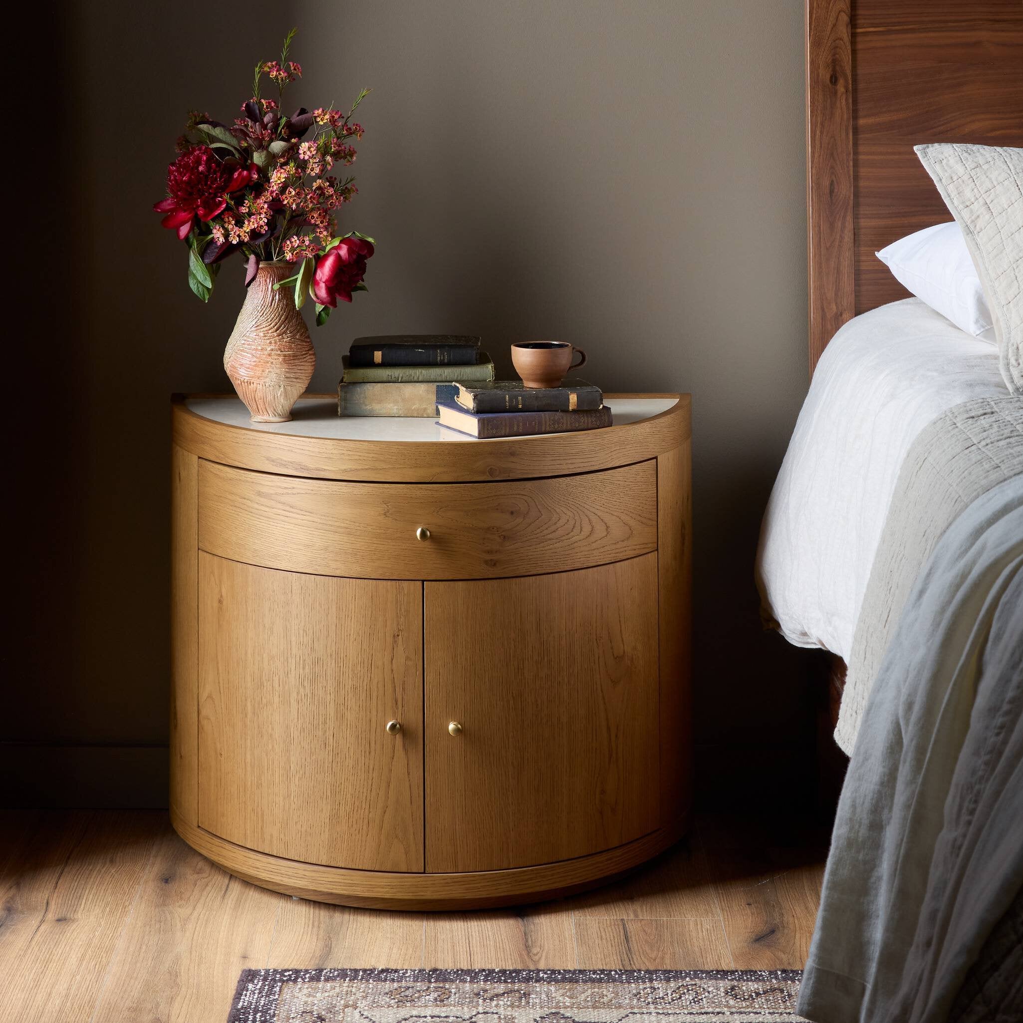 Wooden nightstand with a vase of flowers and books next to a bed.