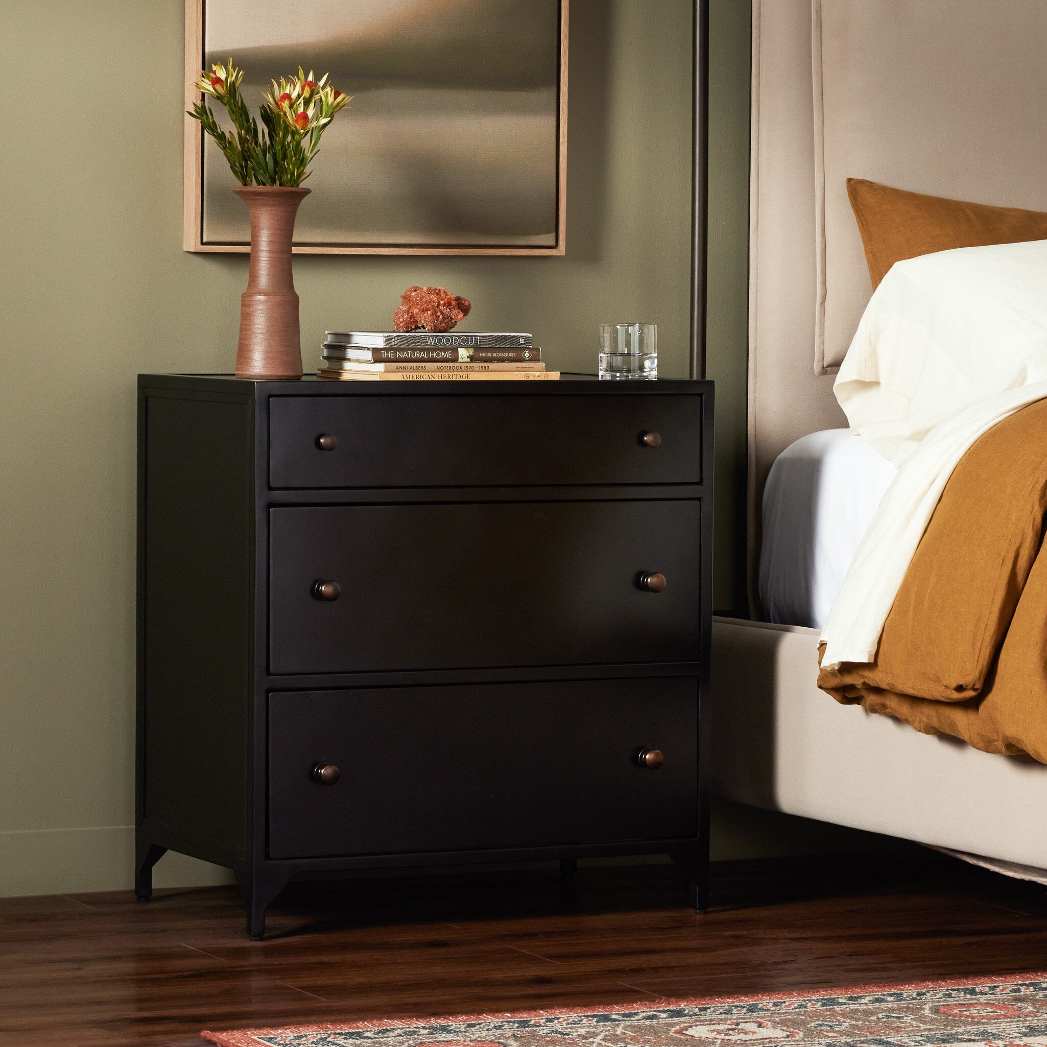 Black dresser next to a bed with a vase and books on top in a bedroom setting.