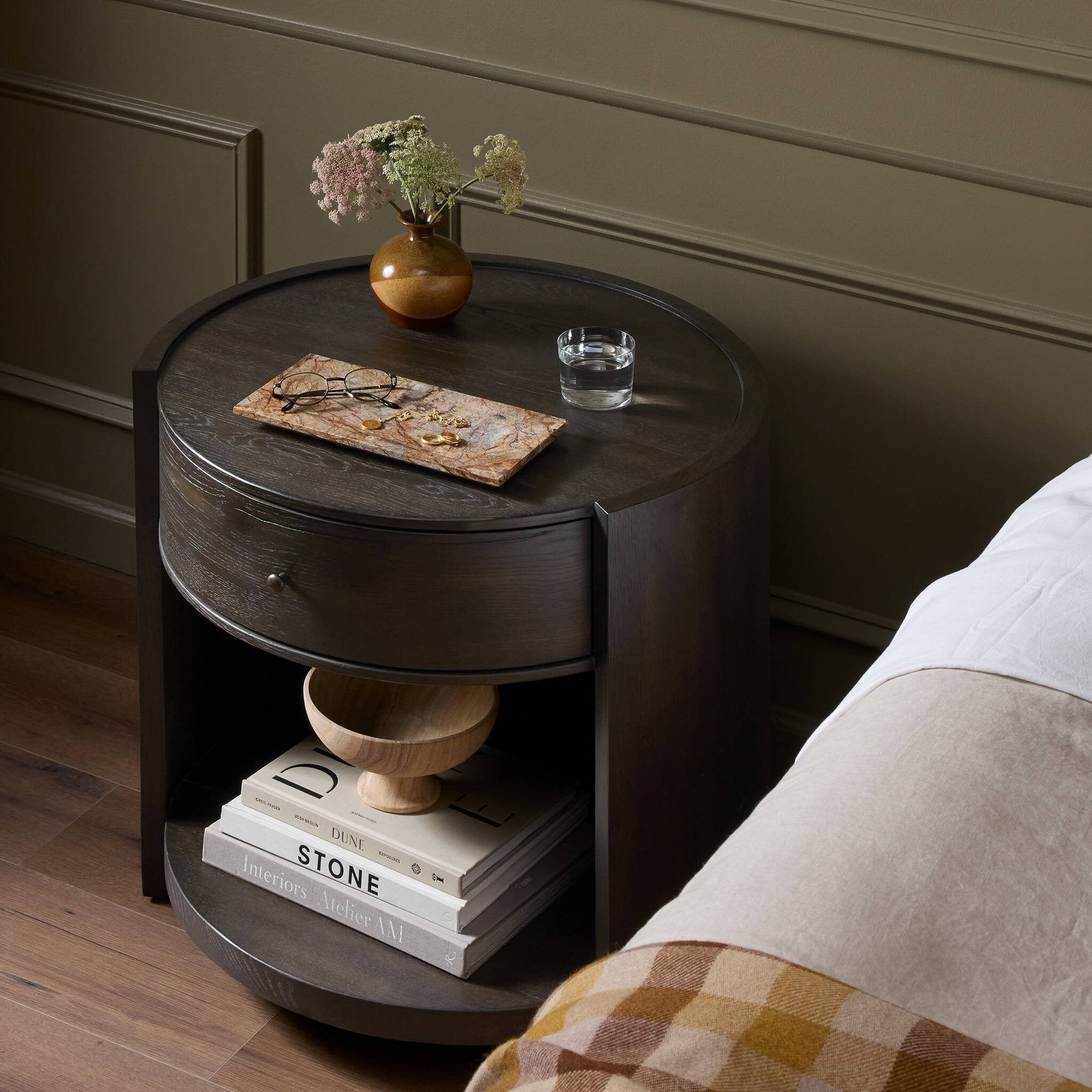 Round wooden nightstand with a drawer, books, and a vase on a wooden floor.