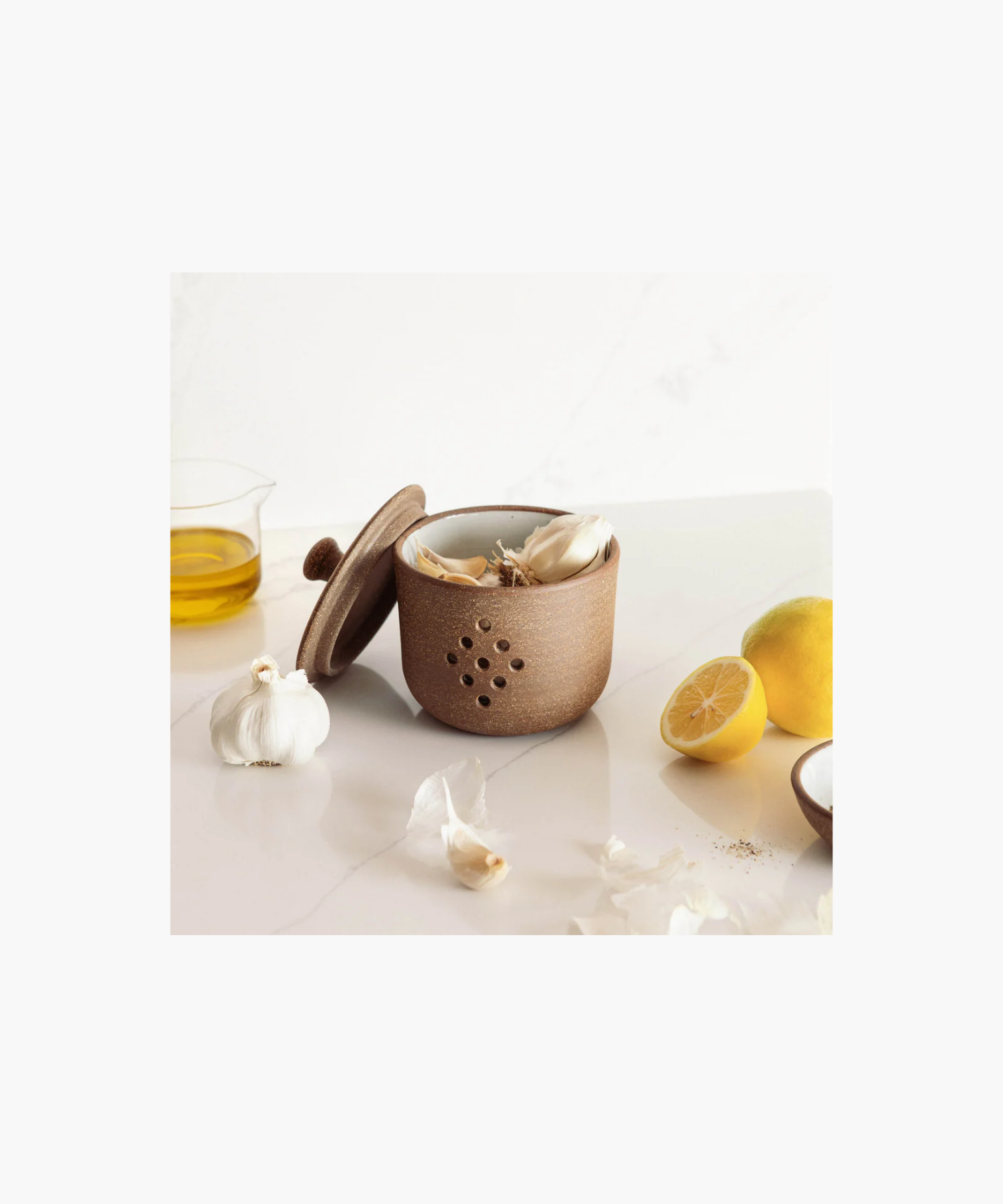 A round, speckled brown pottery garlic keeper with a fitted lid, featuring nine ventilation holes in a diamond pattern. Styled wit garlic on a marble counter. The tone is rustic and earthy.