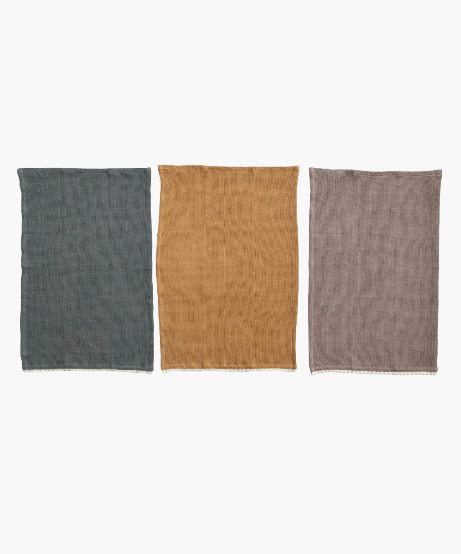 Three unfolded waffle-knit towels in taupe, teal, and mustard are lined side by side. Each towel features a delicate white lace trim along the edge.