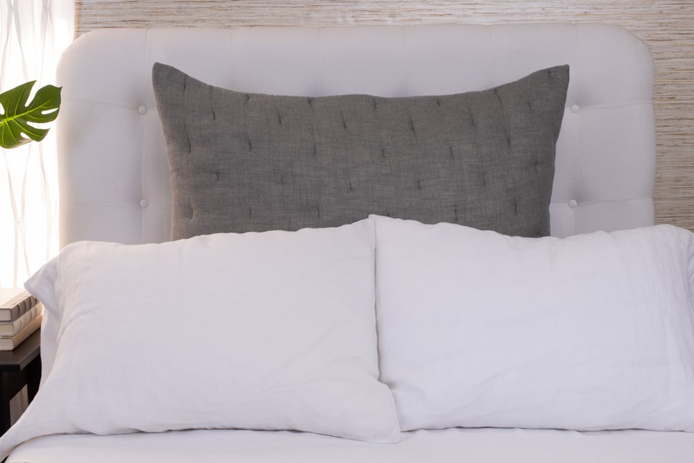 A styled bed with white bedding and a large dark grey pillow on a neutral background.