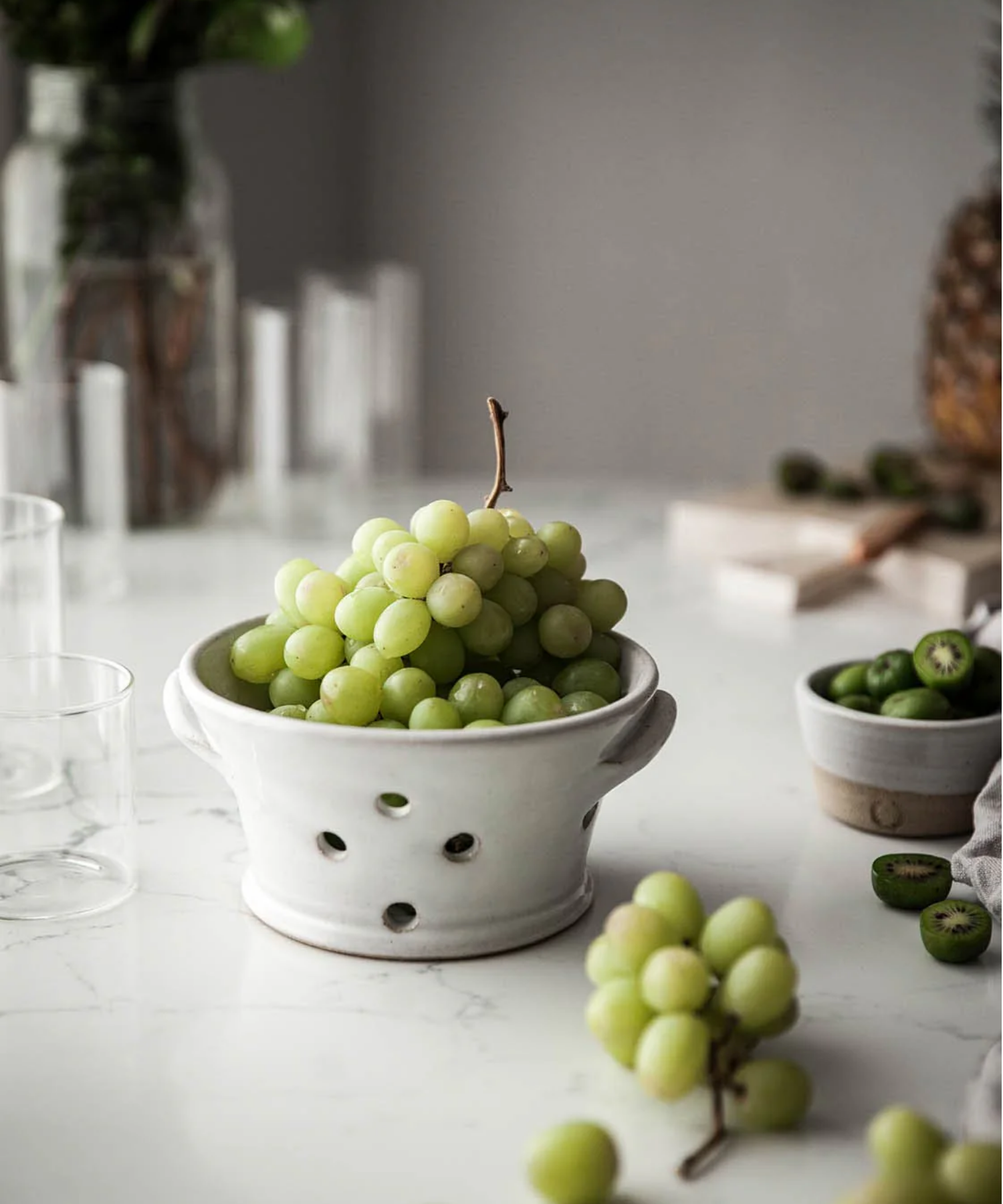 White ceramic colander with two side handles and five round drainage holes styled on a counter with grapes. The smooth, minimalist design conveys a clean and simple aesthetic.
