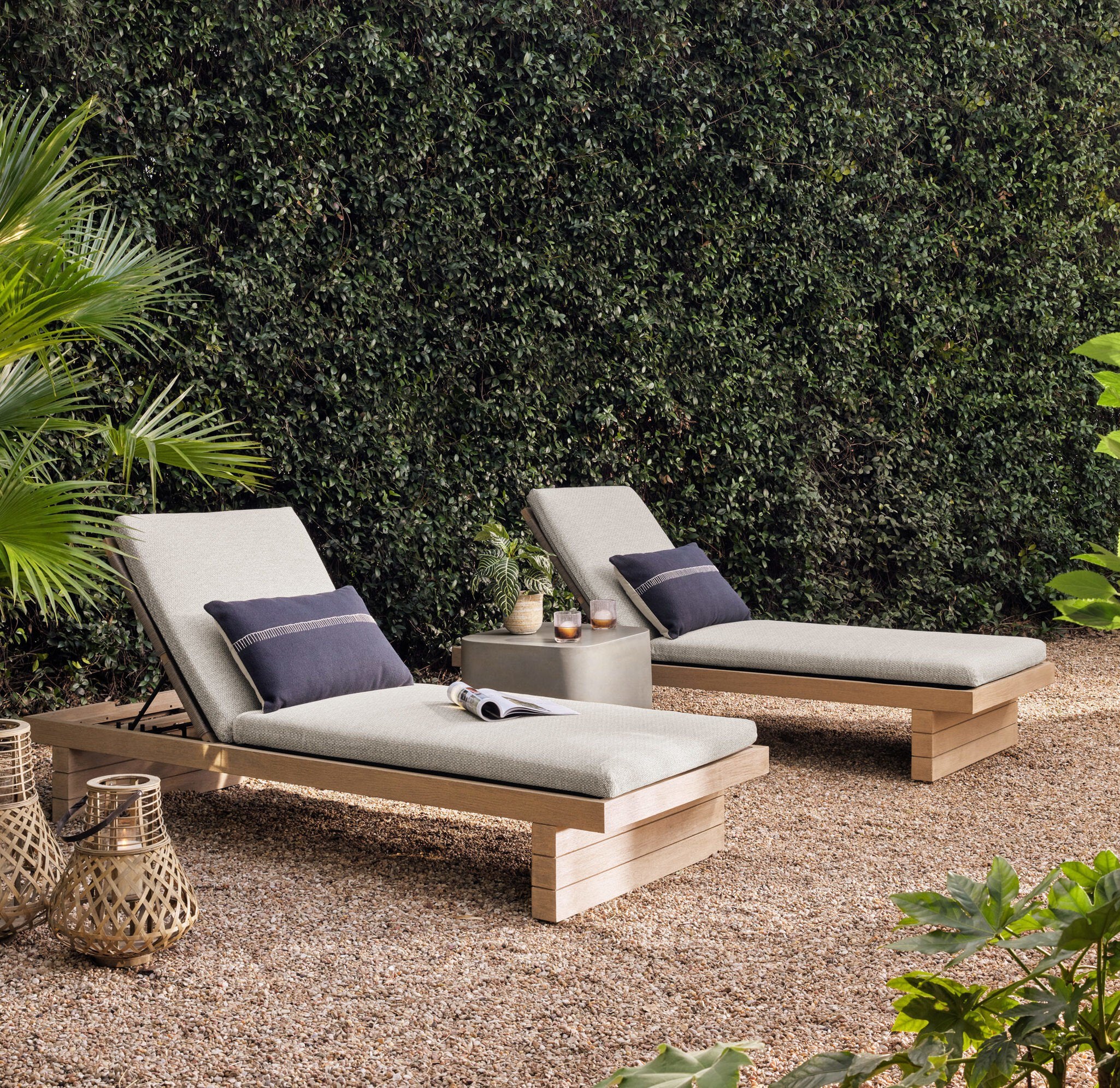 Two outdoor lounge chairs with cushions on a gravel surface with greenery in the background.