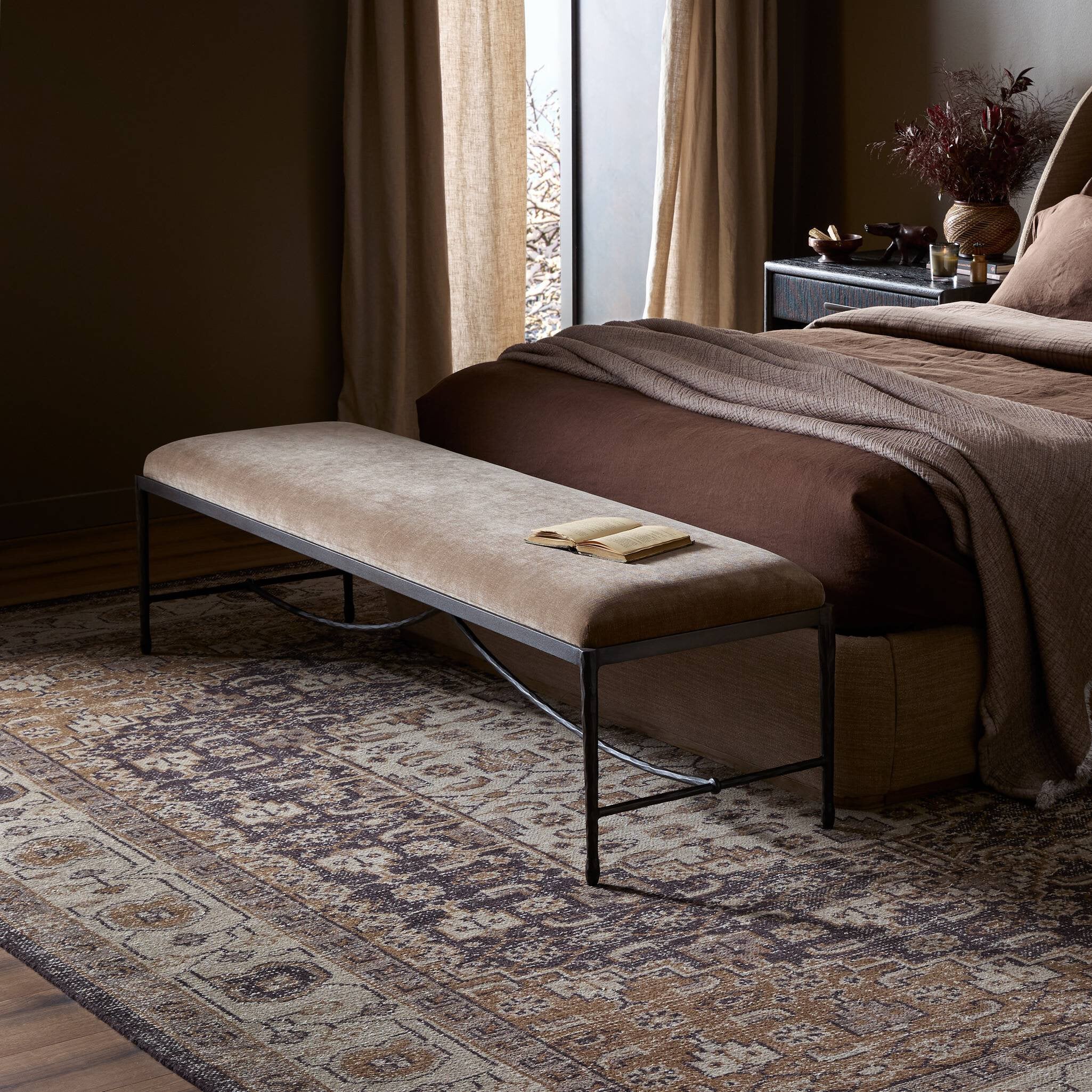 Beige bench with a book on a patterned rug in a bedroom setting.
