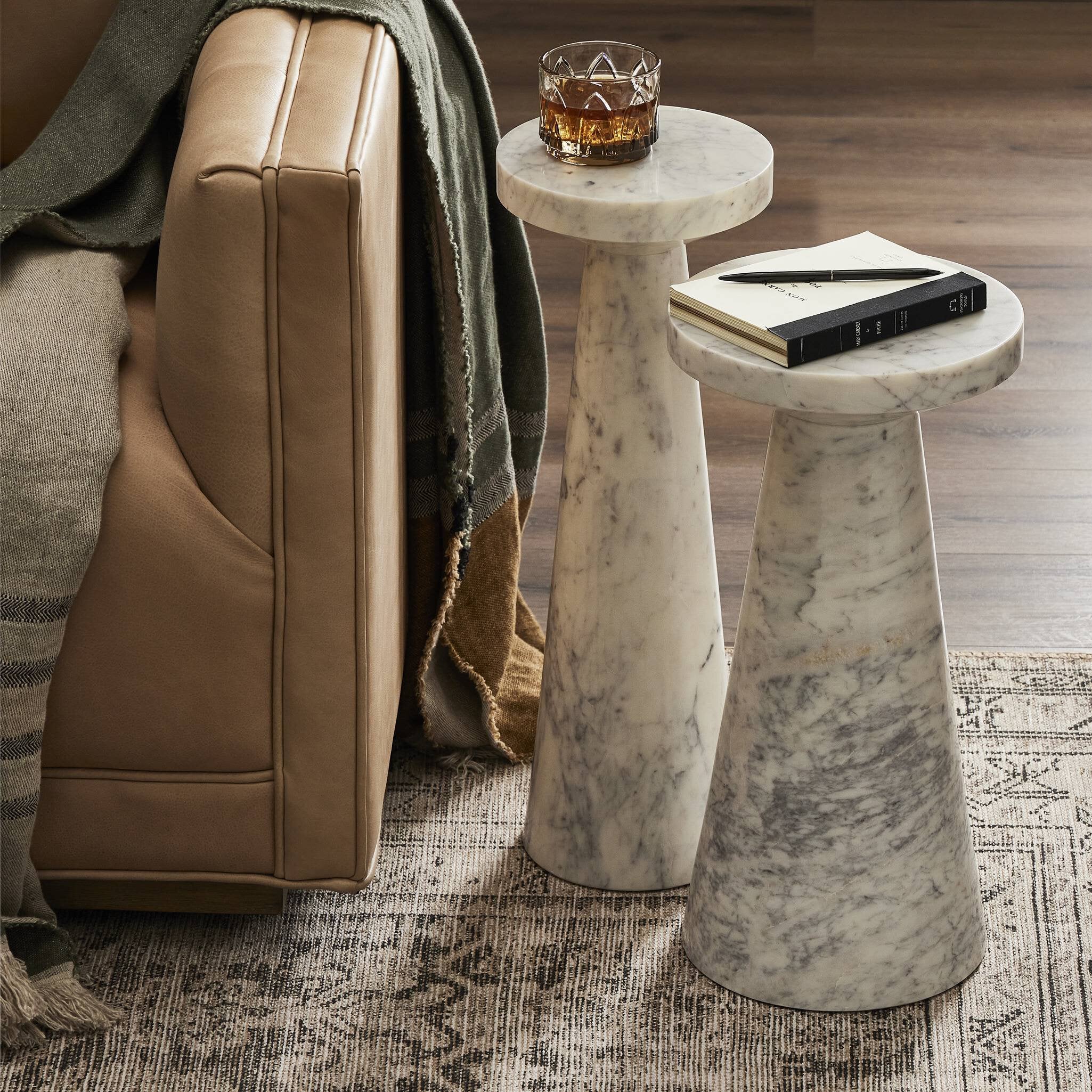 Marble nesting tables with a glass of whiskey and a book next to a brown leather sofa.