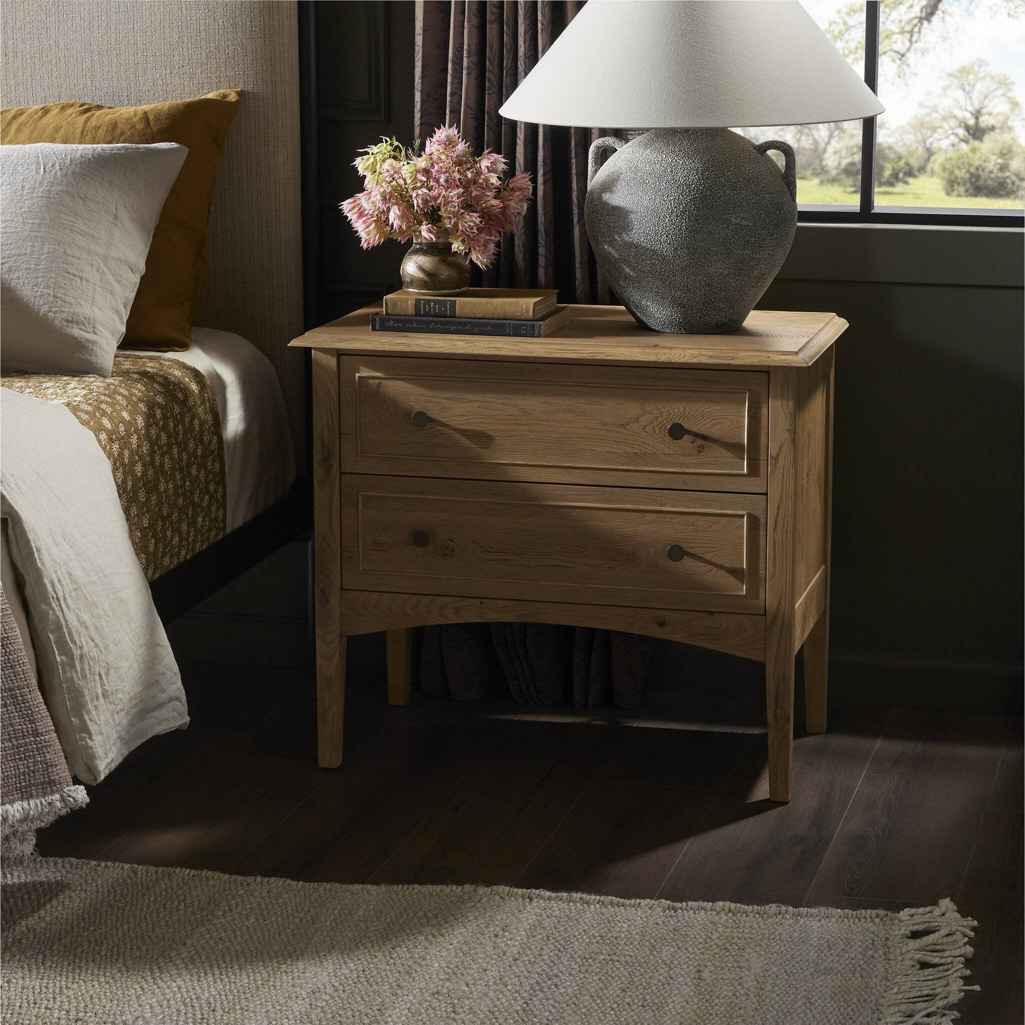 Wooden nightstand with two drawers styled in a bedroom with a lamp and books.