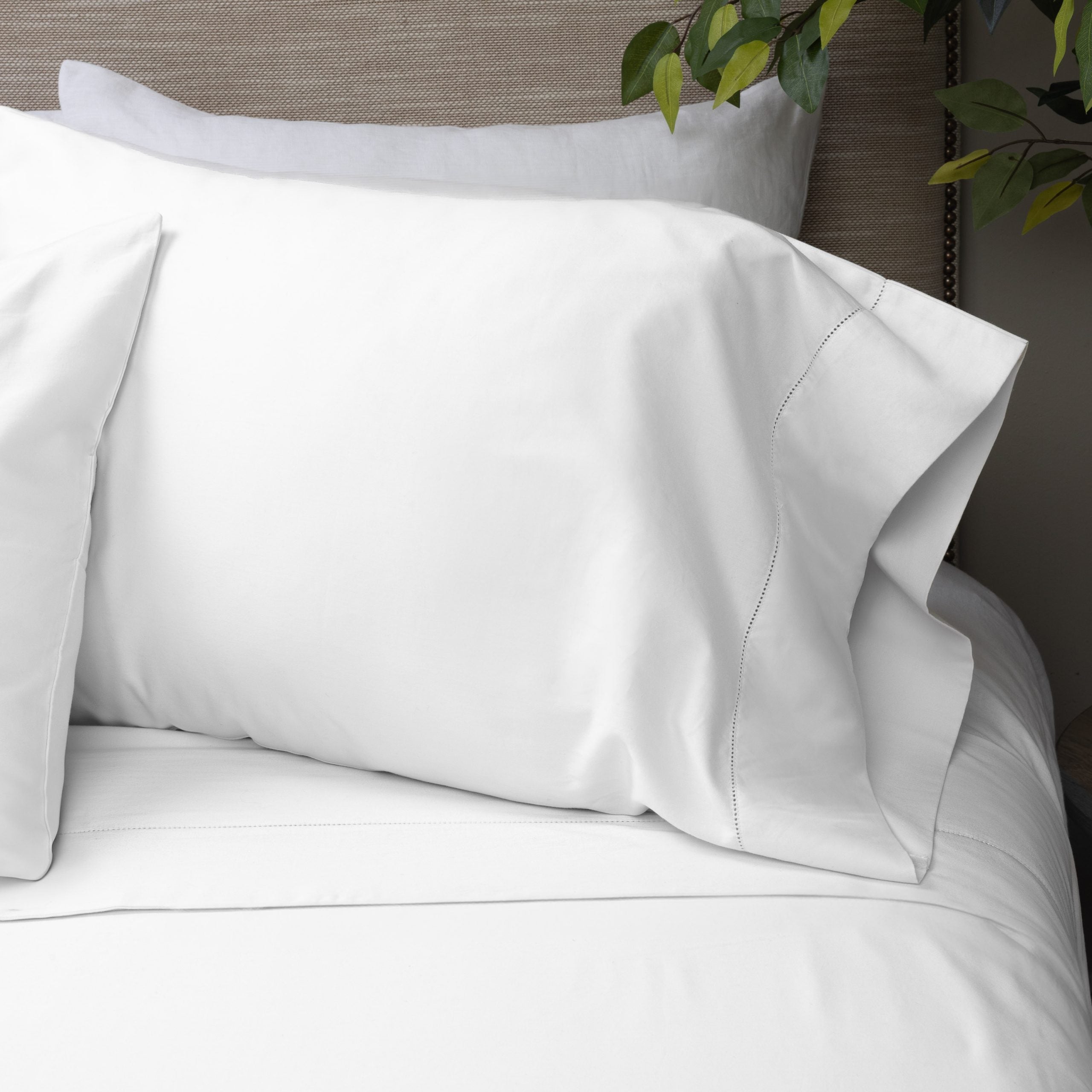 Close-up of a styled bed featuring the Murano white pillowcase with a hemstitch border, placed on a bed with neutral bedding and greenery in the background.