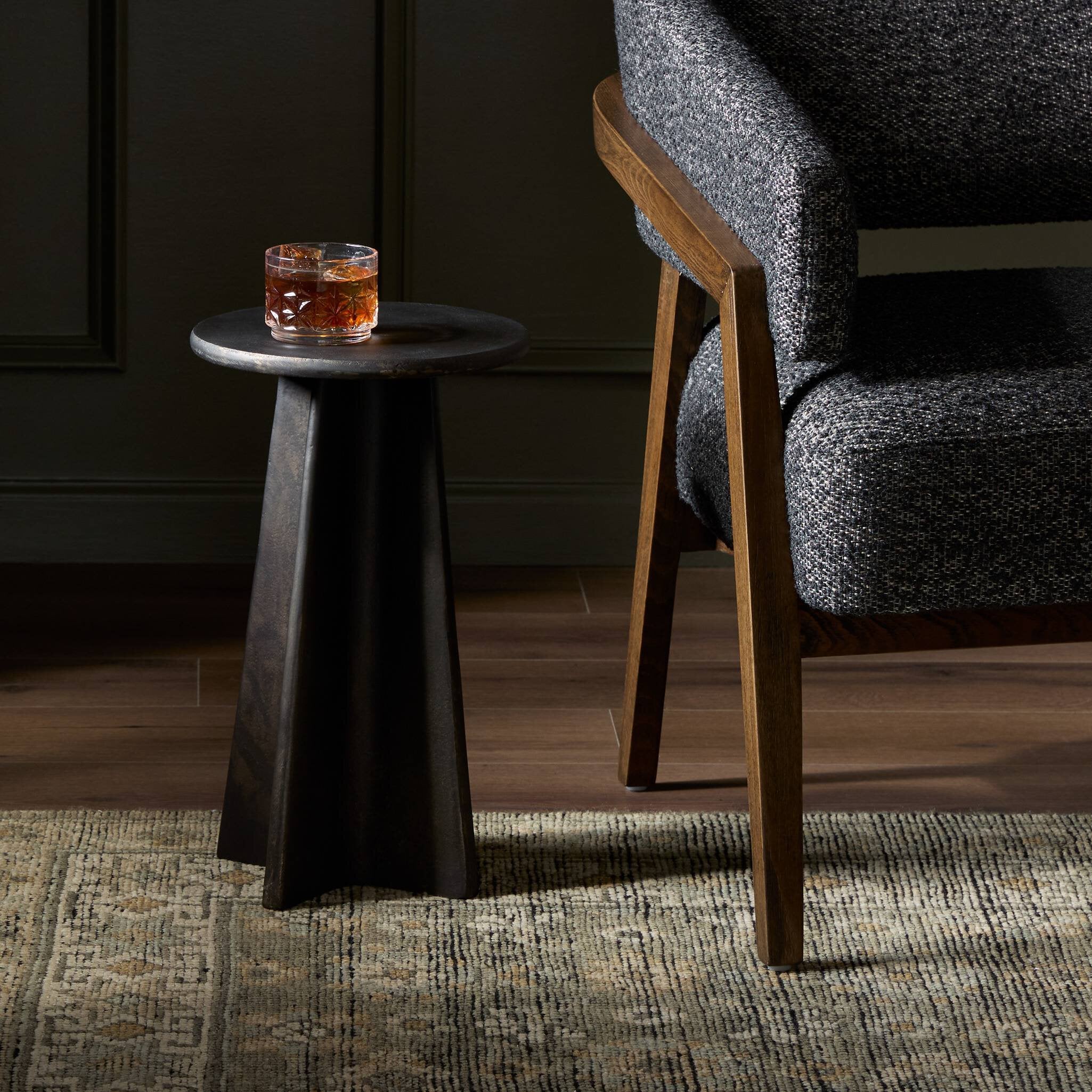 Small round distressed bronze side table with a glass of whiskey next to a gray upholstered chair.