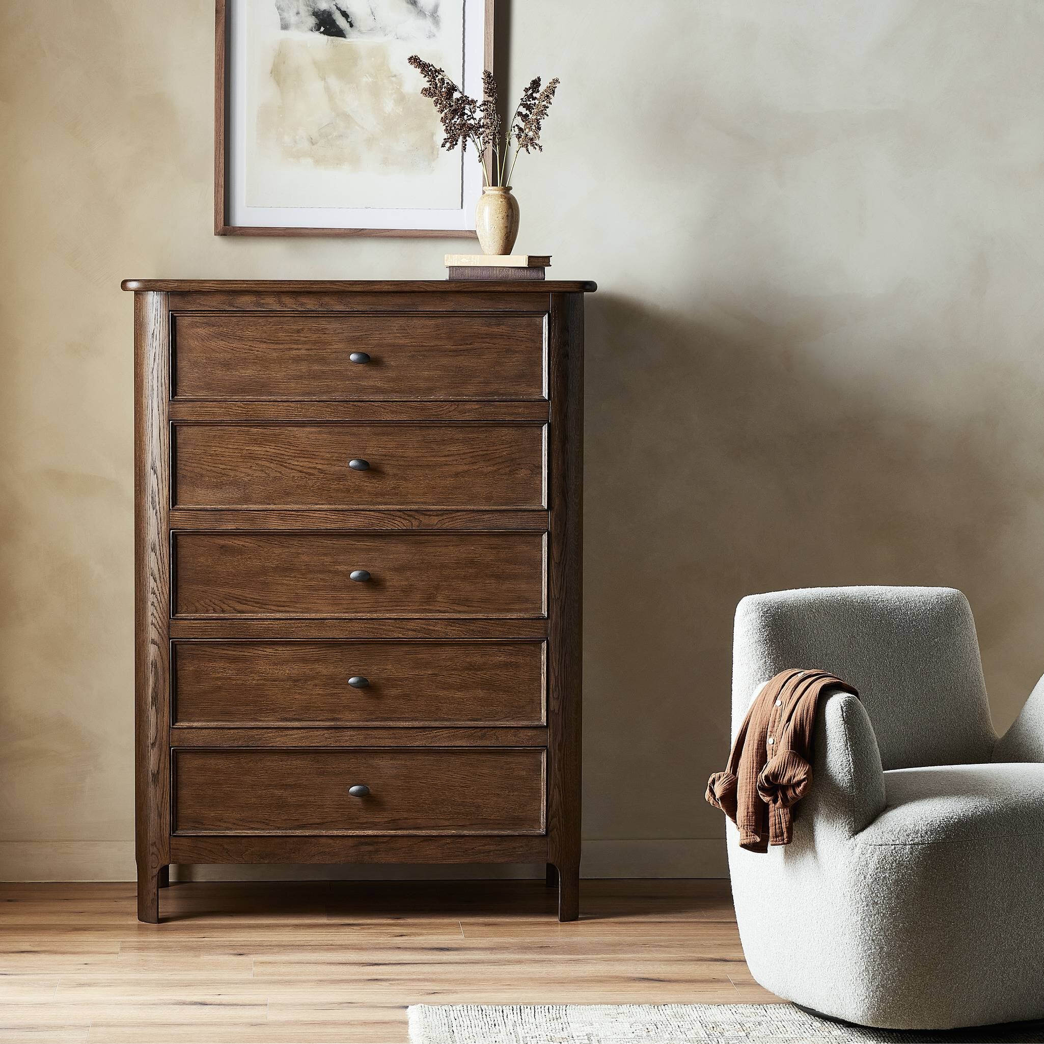 Wooden dresser with five drawers next to a gray armchair in a room with a neutral wall.