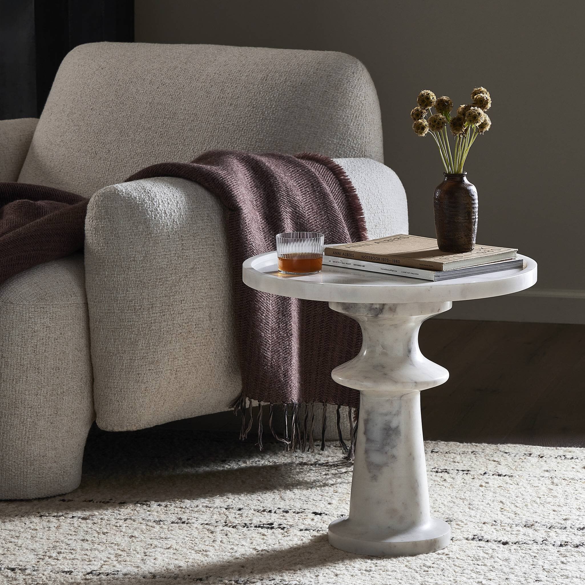 White marble side table with a glass of whiskey and a book next to a beige armchair with a brown blanket.