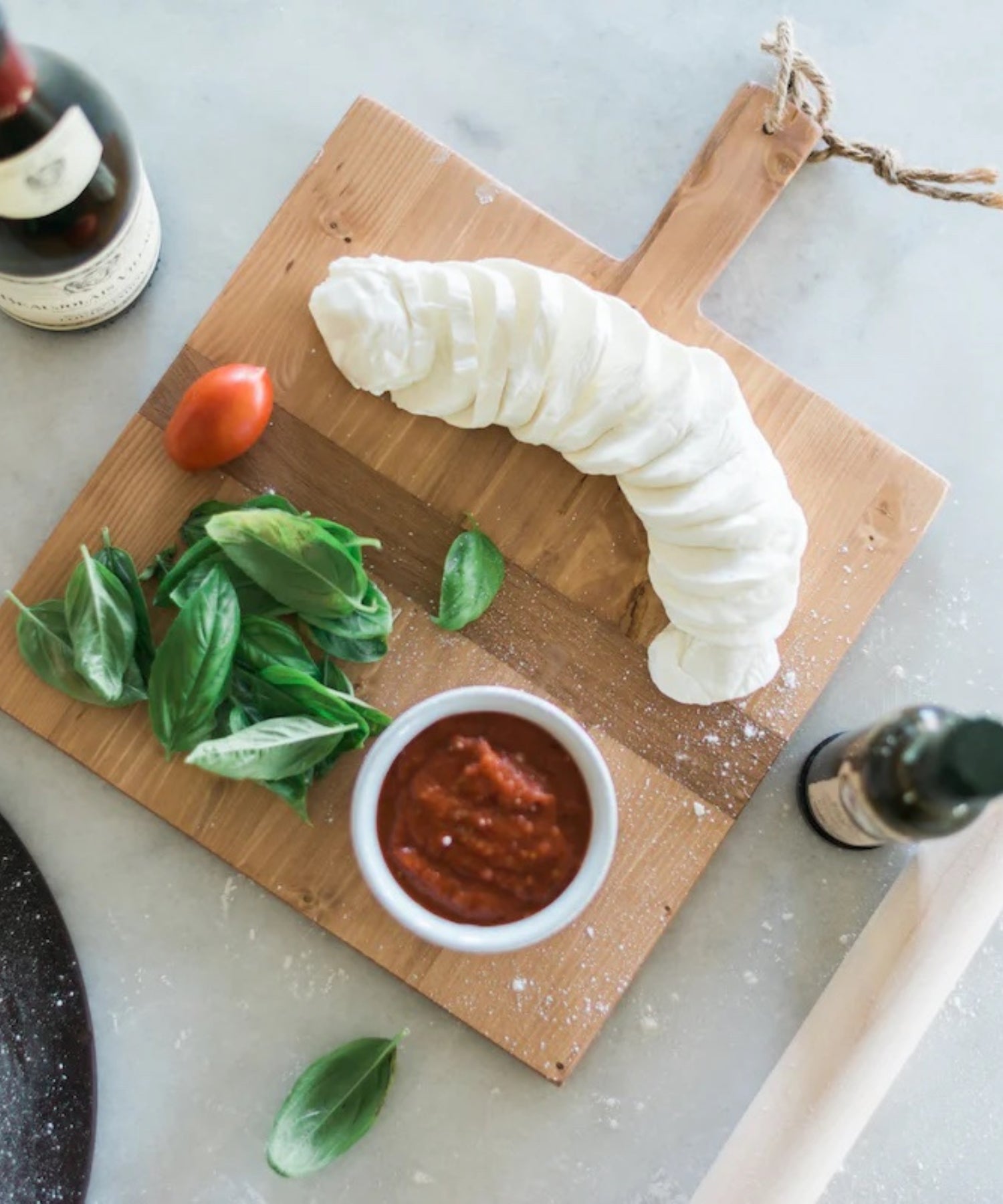 Square pine wood cutting or charcuterie board with a natural finish, featuring a dark horizontal stripe and a handle with a rope loop styled with cheese and wine.