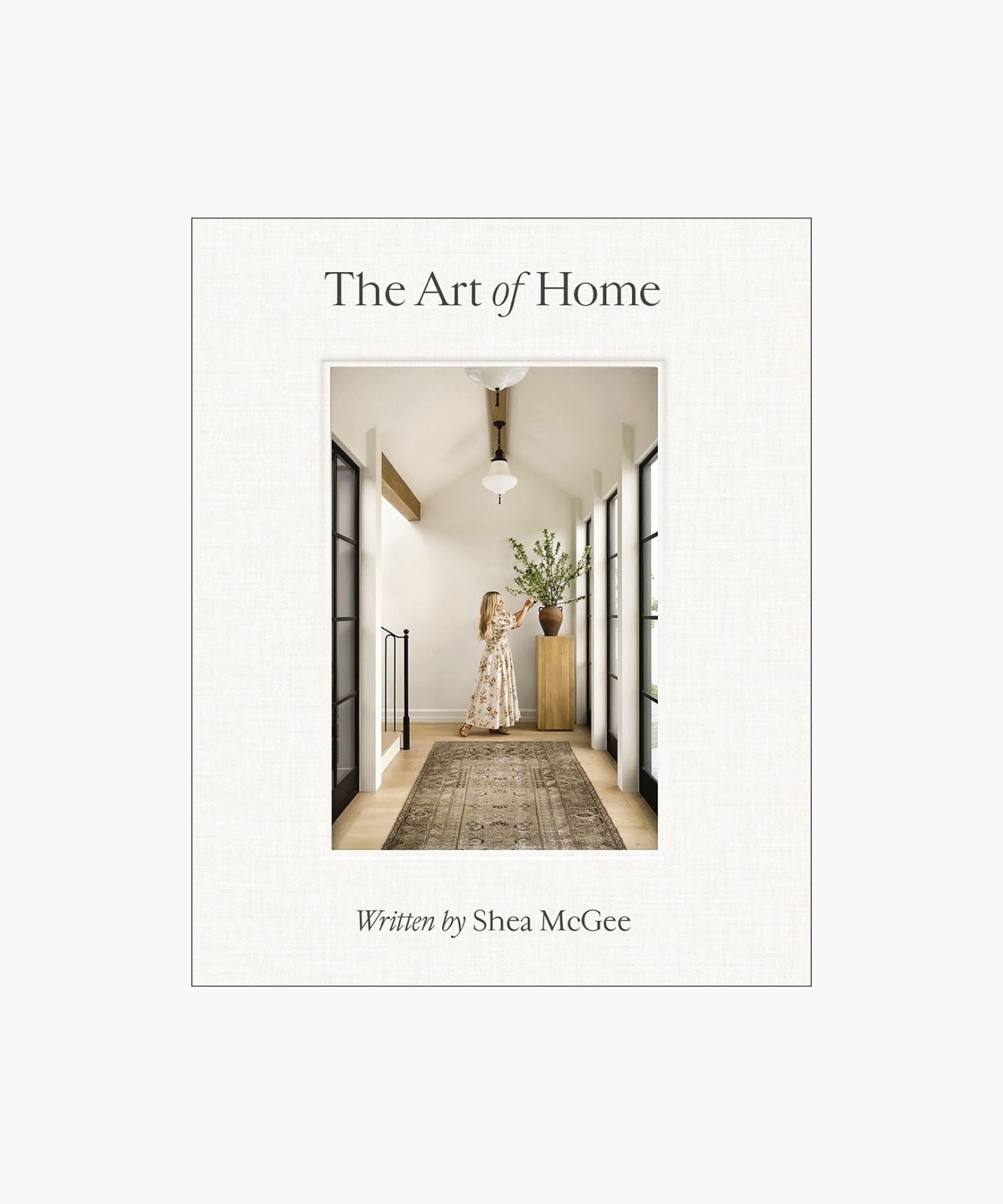The cover of the book "The Art of Home" by Shea McGee features a woman arranges branches in a vase on a wooden stand, in a bright hallway with large windows and a patterned rug.