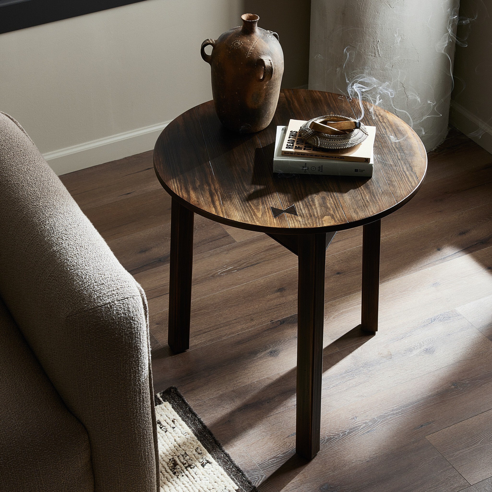 Round wooden end table with a vase and books in a room with a sofa and rug.