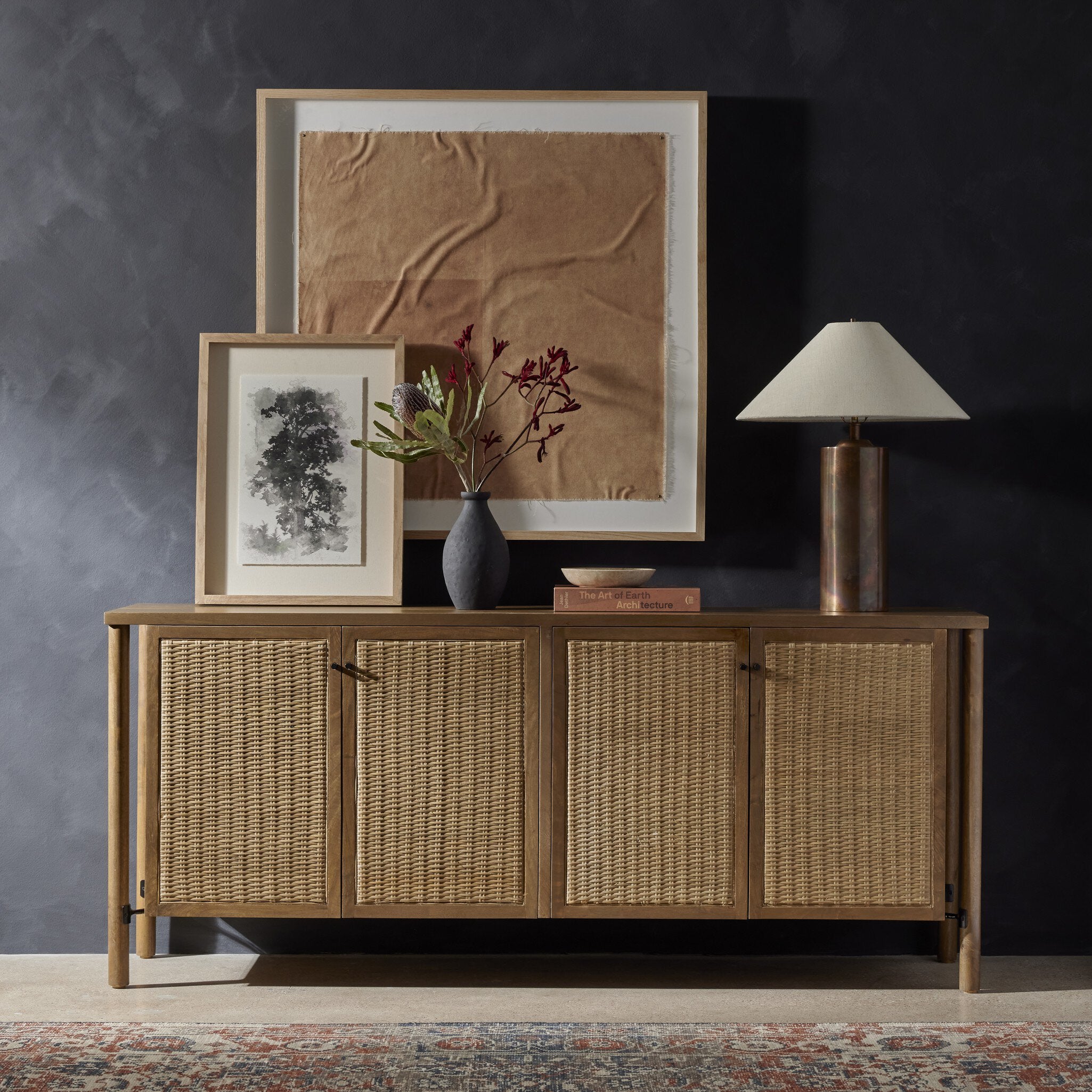 Wooden sideboard with wicker doors, framed artwork, and a lamp against a dark wall.