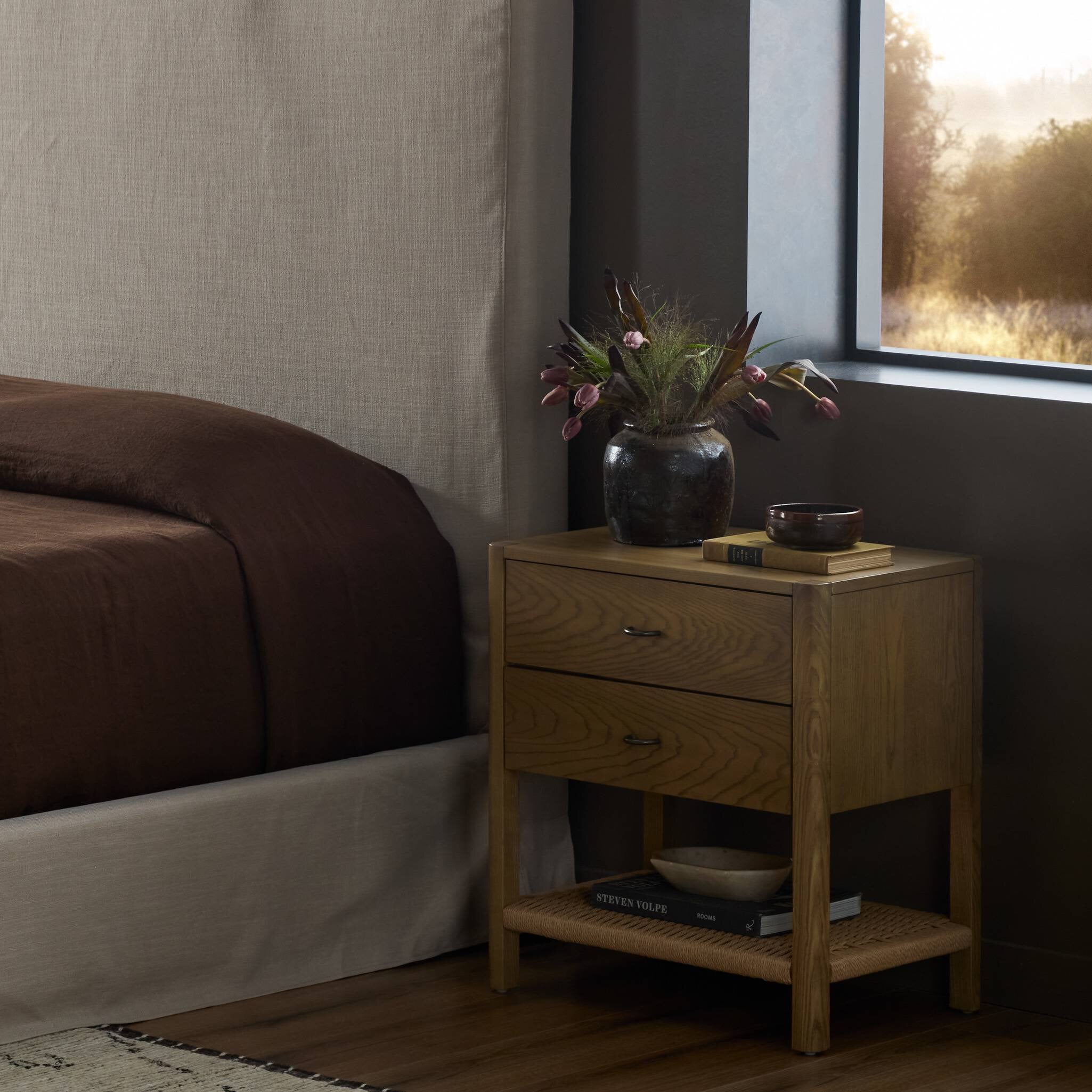 Wooden nightstand with a vase of flowers next to a bed in a bedroom.