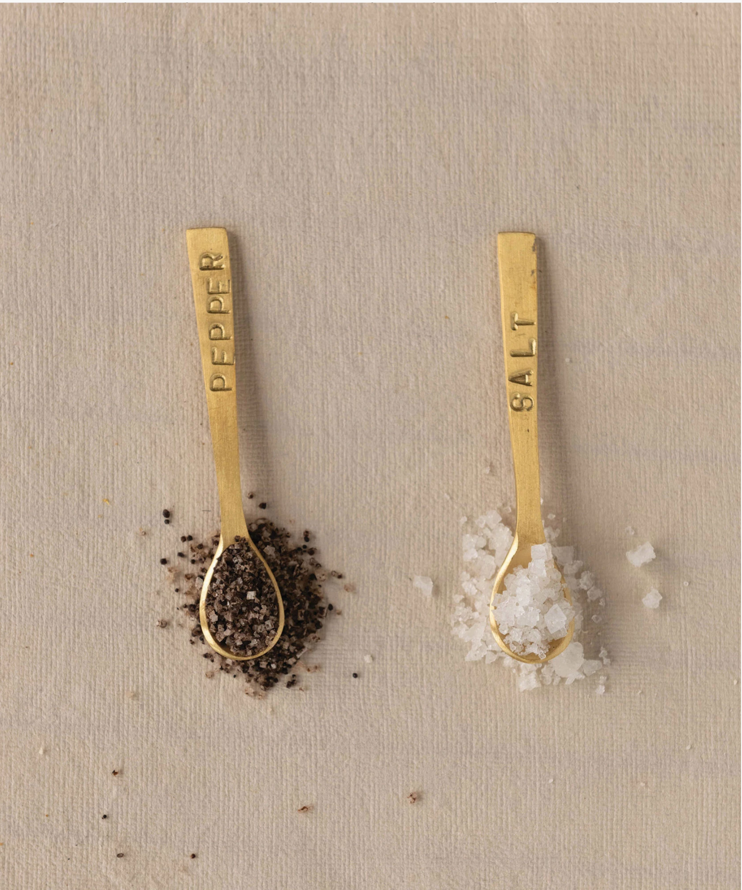 Two small elegant gold-toned spoons with engraved labels, "SALT" and "PEPPER," on their handles styled each with salt and pepper set against a neutral background, conveying sophistication.