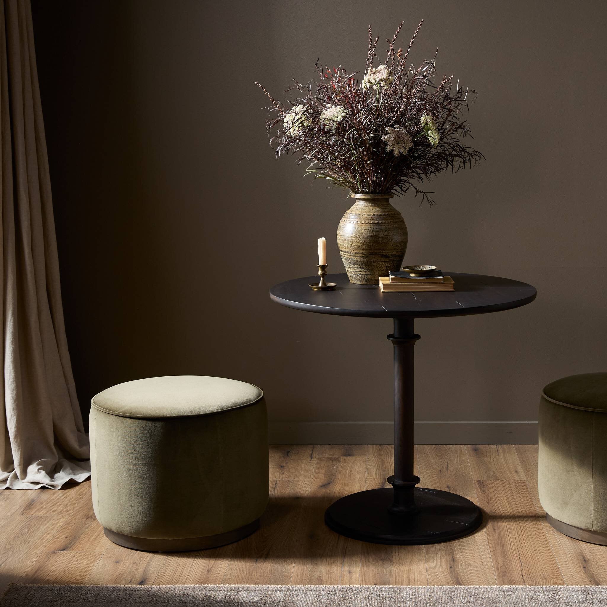 Small round table with a vase of flowers and books against a dark wall, next to a cushioned ottoman.