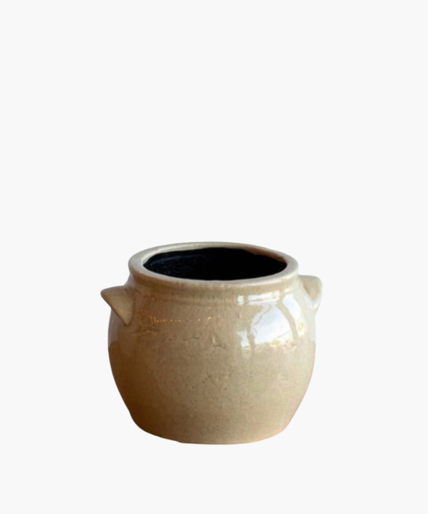 A beige ceramic pot with a glossy finish, featuring two small side handles and a black interior. The pot has a smooth, rounded shape and exudes a rustic charm.