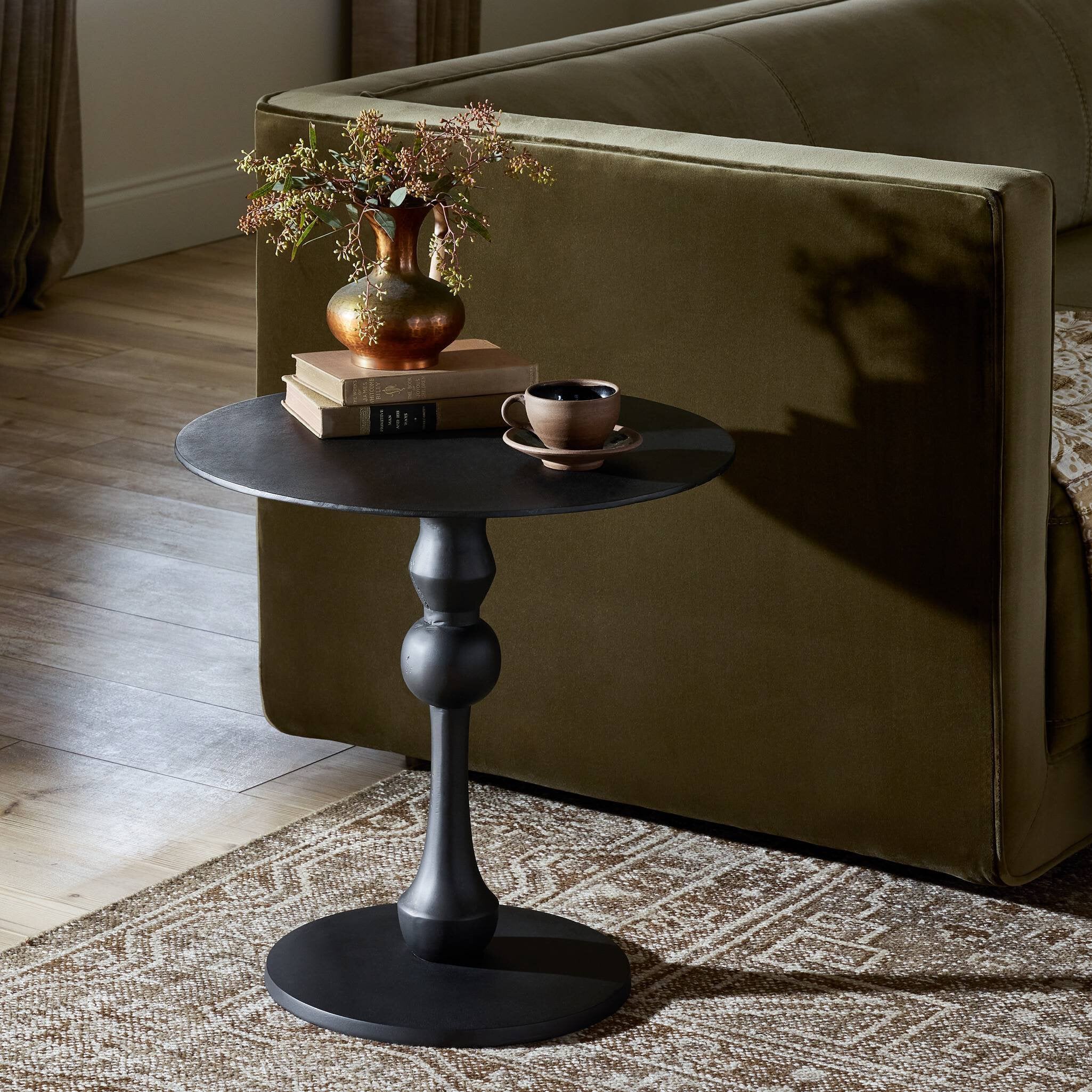 Small black side table with a vase, books, and cup in front of a beige sofa.