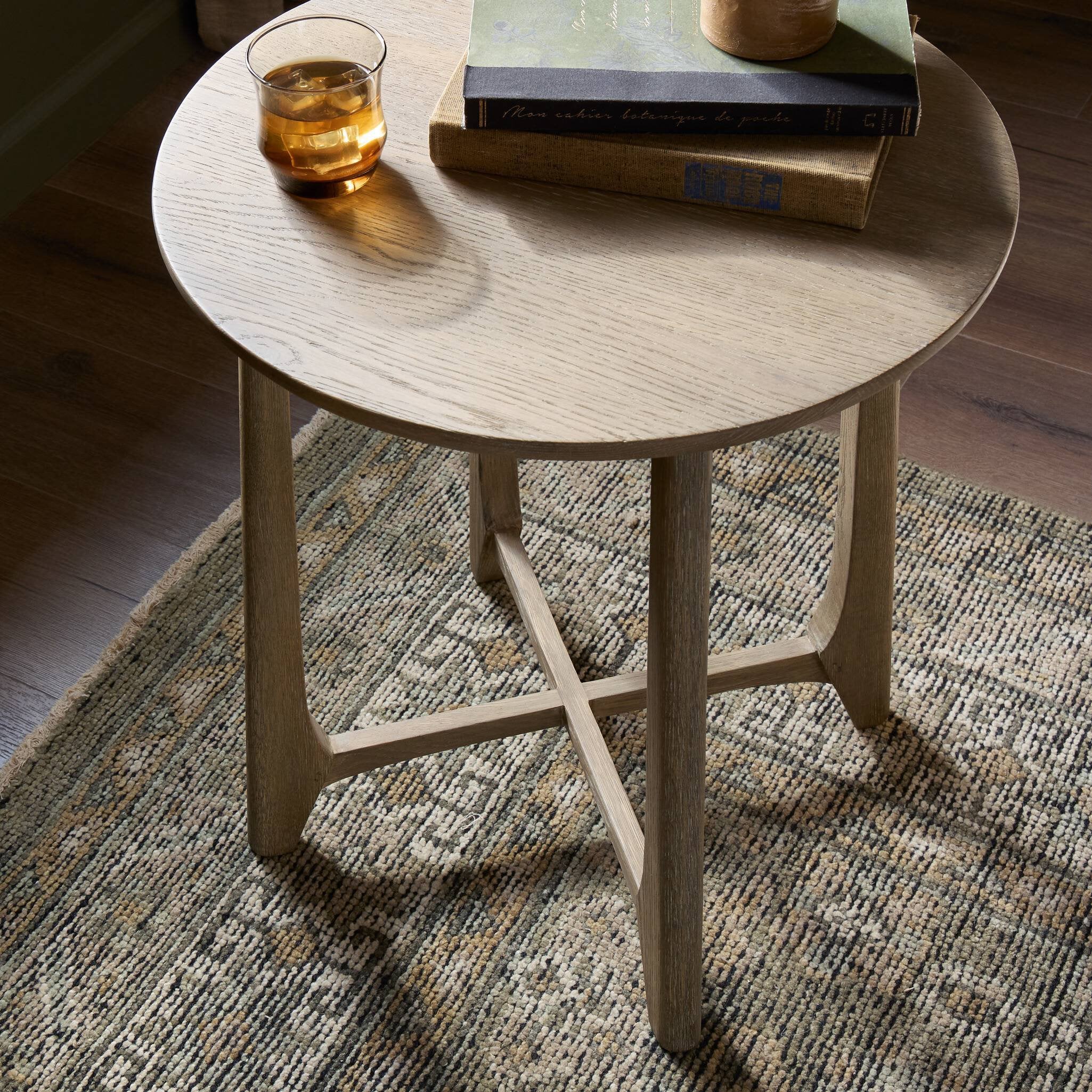 Round wooden side table with a glass of amber liquid and books on a patterned rug.