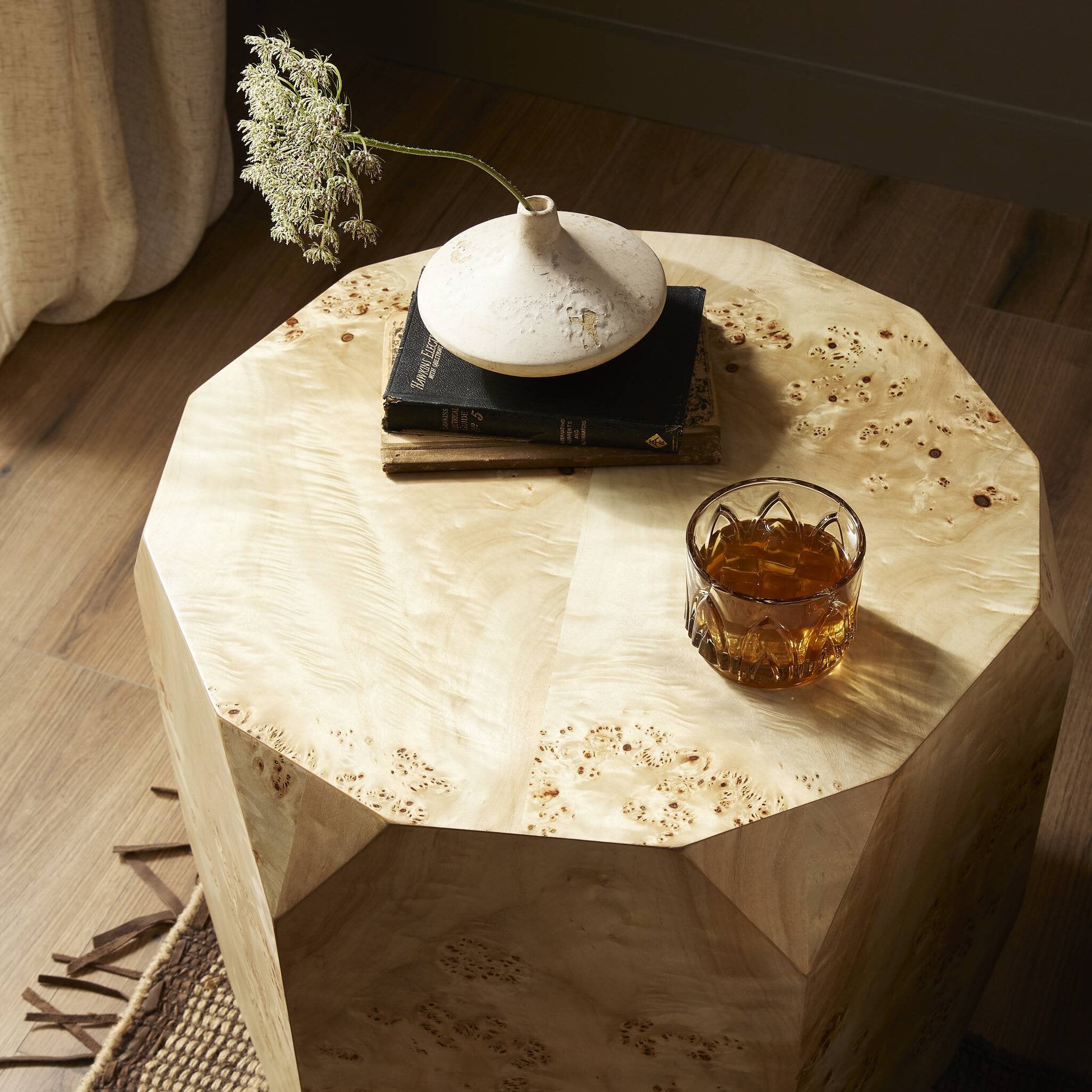 Hexagonal wooden end table with a vase, books, and a glass of whiskey in a warm indoor setting.