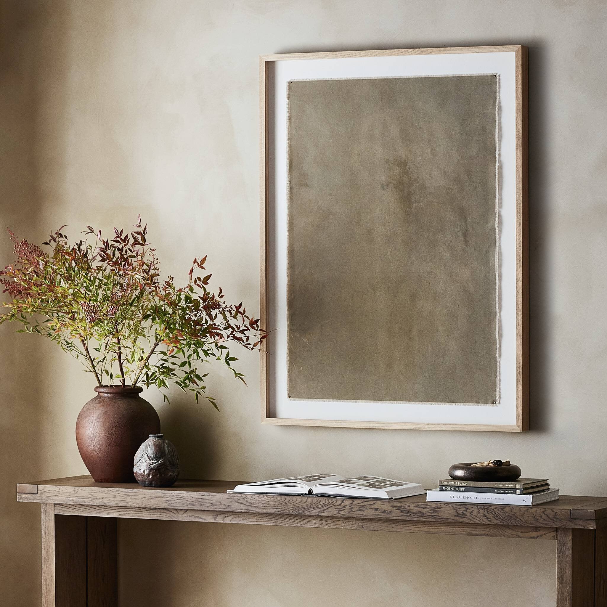 Decorative setup with a vase of flowers, books, and a framed abstract painting on a wooden console against a beige wall.