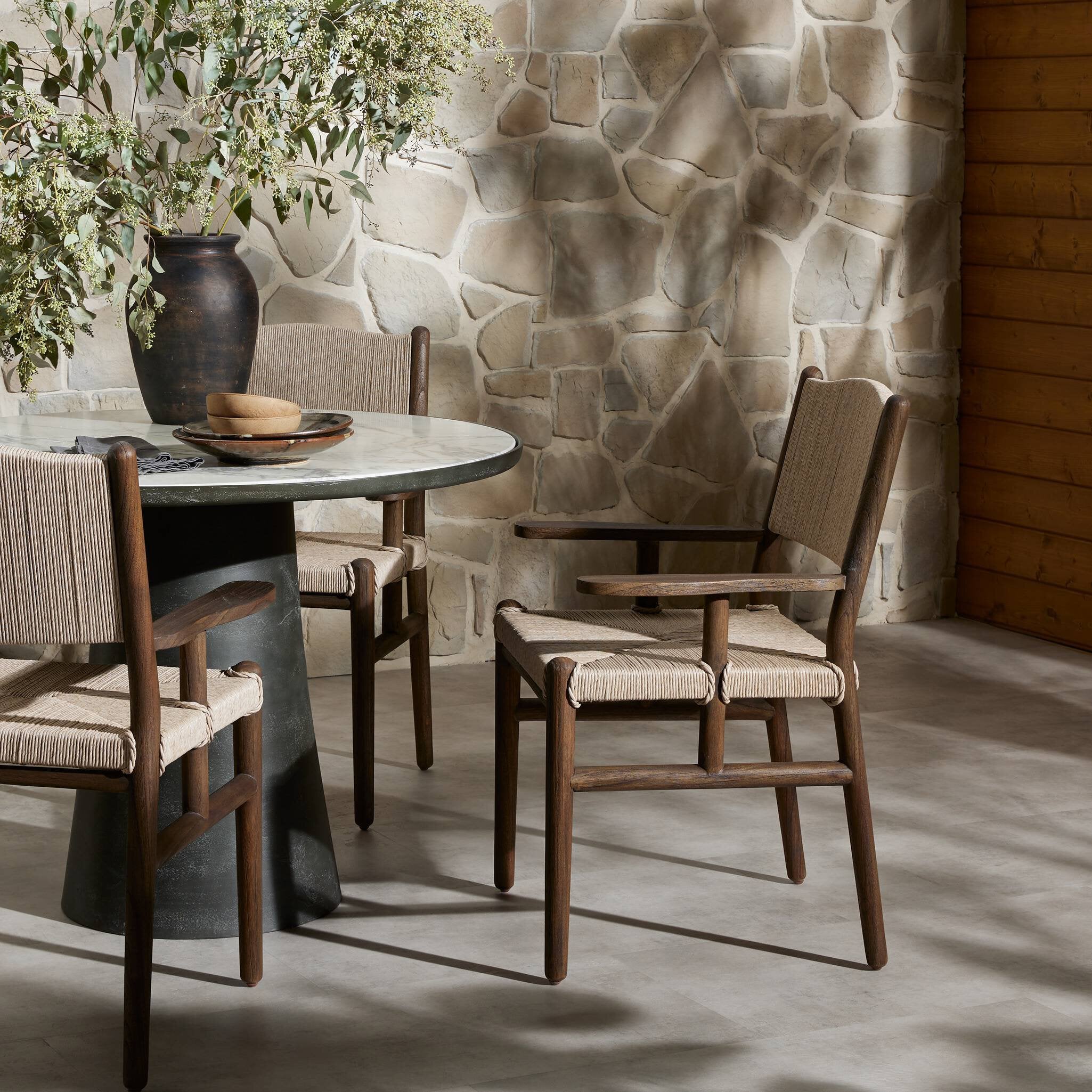 Outdoor dining area with round table and wooden armchairs against a stone wall.