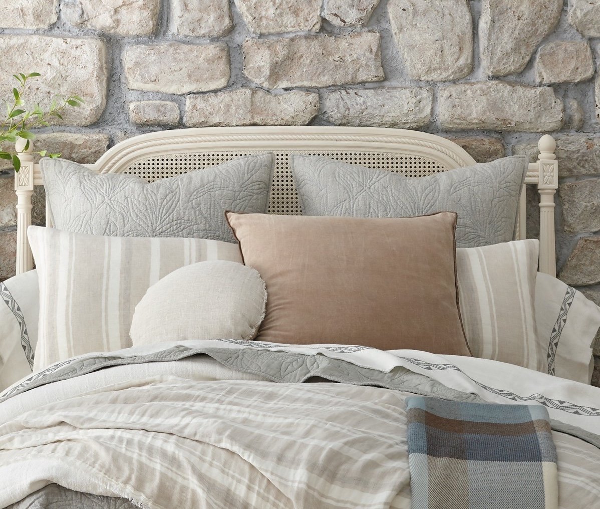 Bed styled with Hopkins shams and coverlet in a layered look, framed by a stone wall and cane headboard for a rustic, cozy bedroom setup.