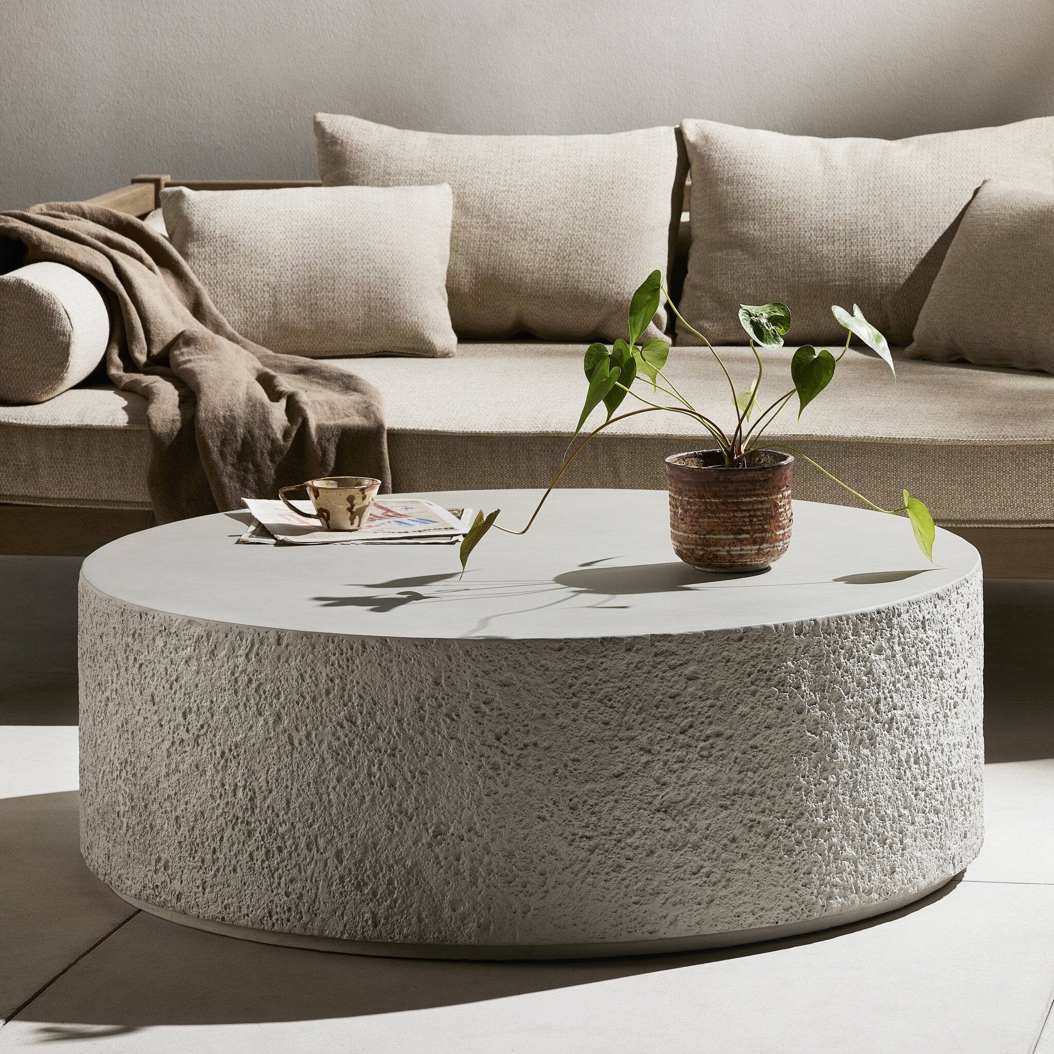 Round concrete coffee table with a plant and cup in front of a beige sectional sofa.