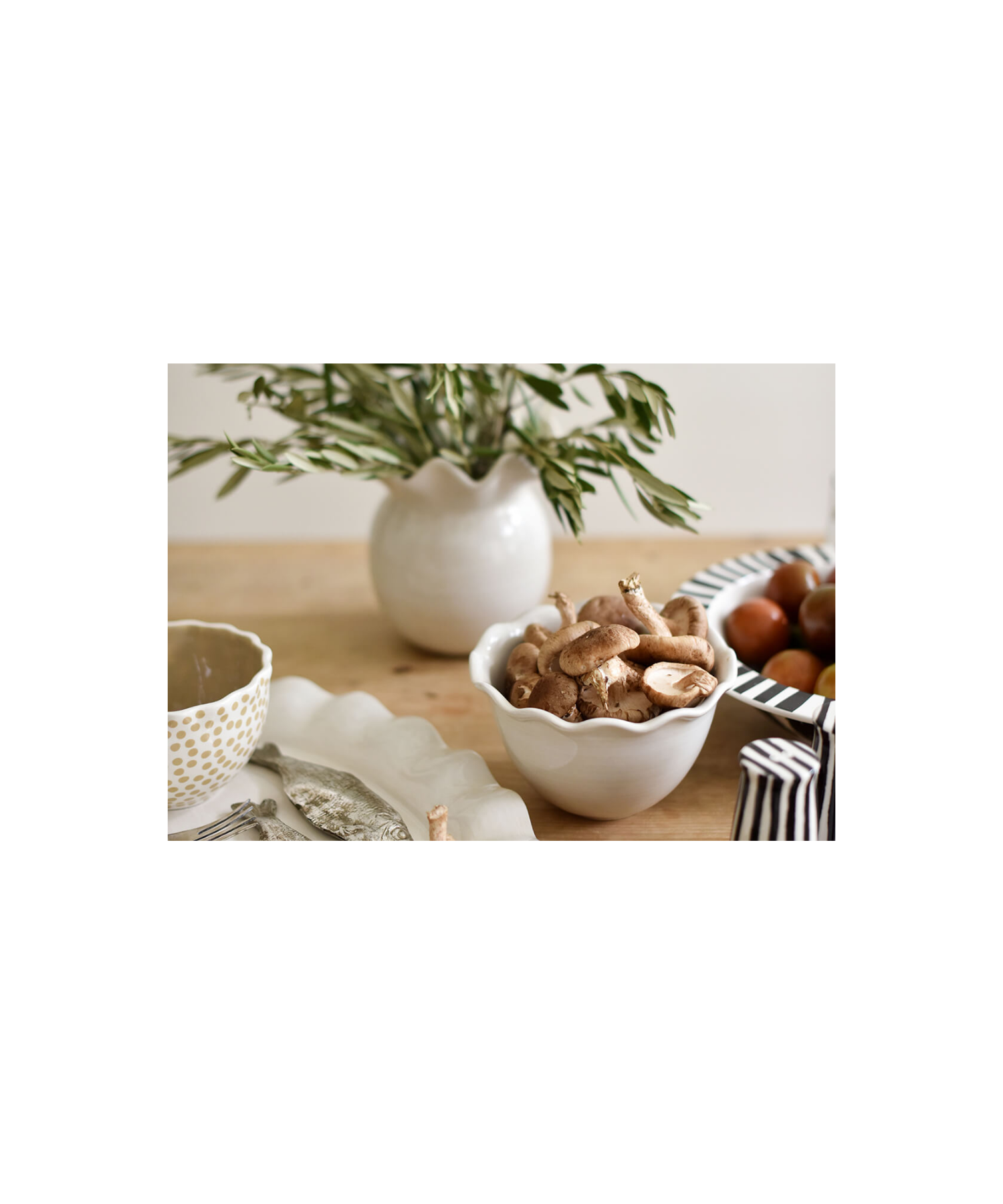 White ceramic vase with a glossy finish and wavy, ruffled rim styled on a table with greenery. The vase has a round, bulbous body, conveying elegance and simplicity.