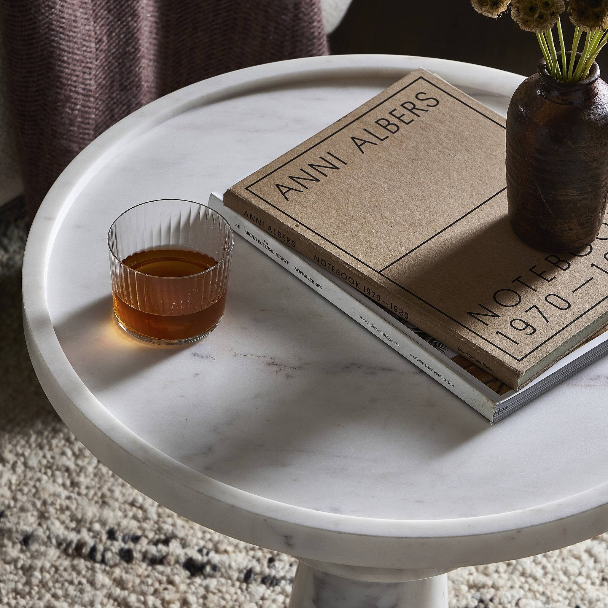 Round marble table with a book, glass of amber liquid, and vase on a carpeted floor.