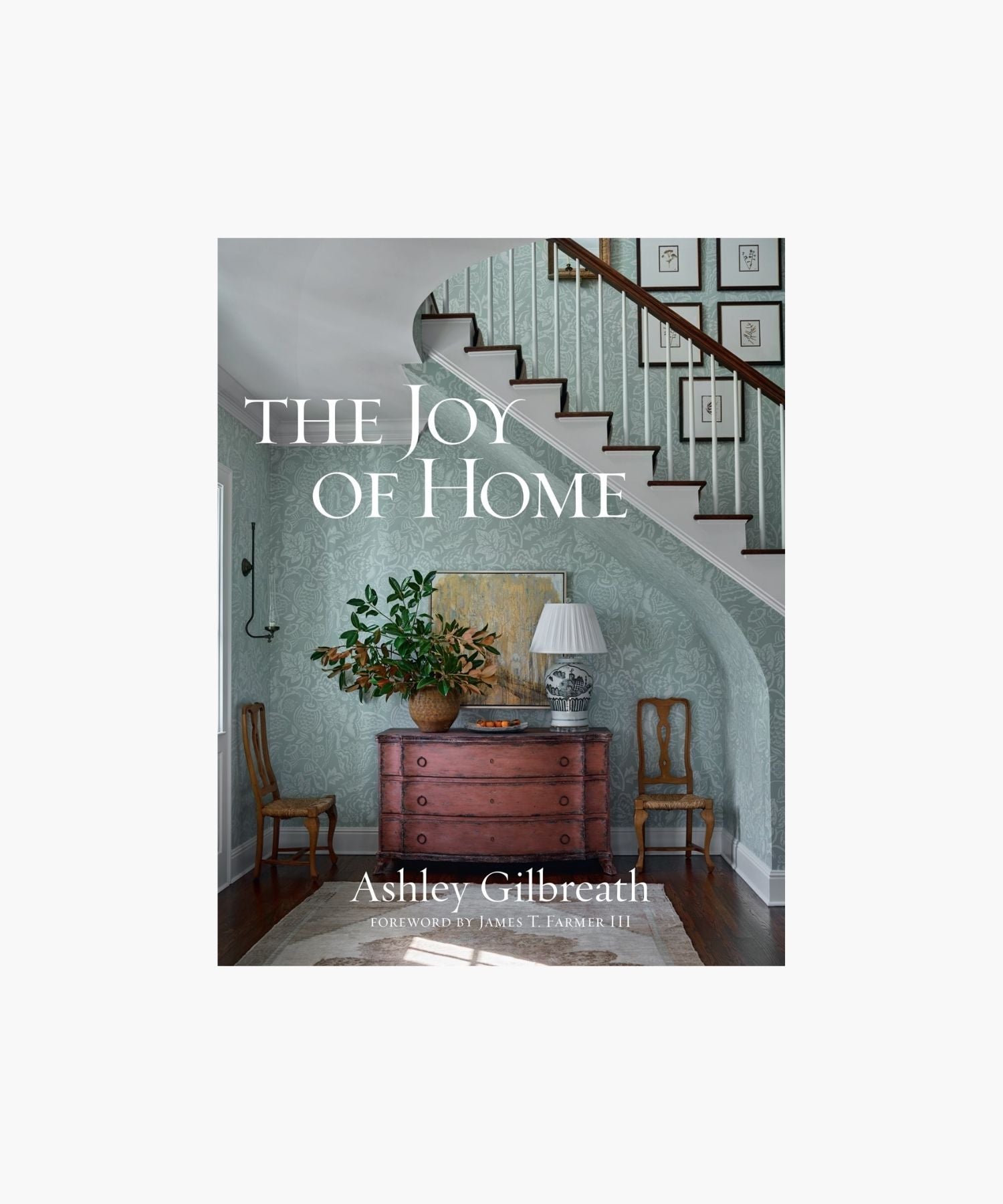 The cover of the book "The Joy of Home" by Ashley Gilbreath features a cozy, elegant hallway with a wooden chest, potted plant, and lamp beneath a staircase; framed prints on the staircase wall. Classic and inviting.
