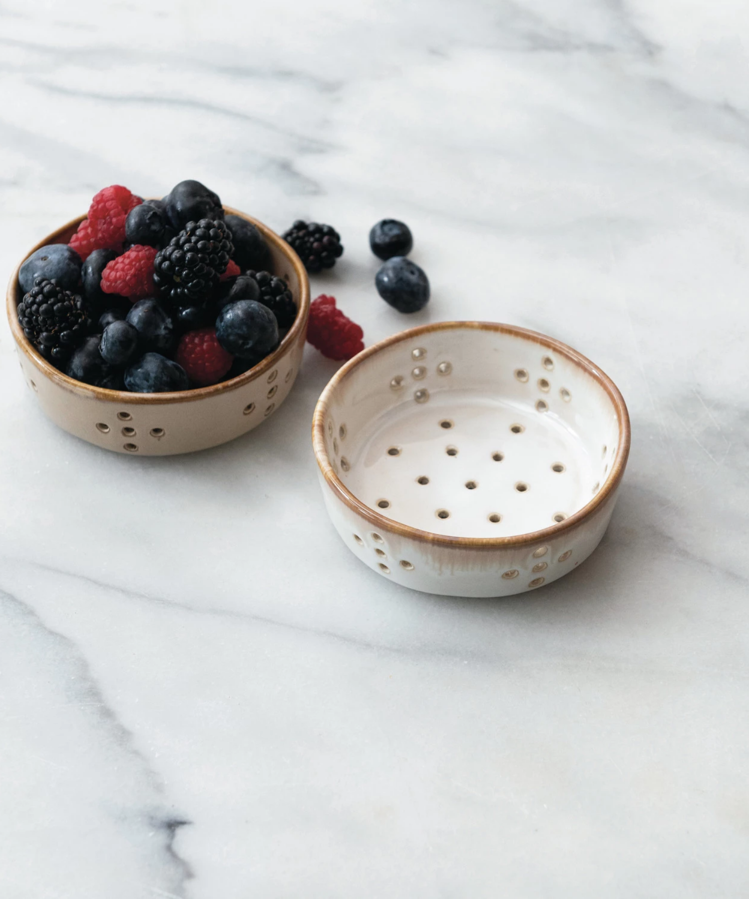 A beige and white ceramic colander features multiple circular drainage holes arranged in patterns styled with mixed berries. The bowl has a smooth, glazed finish, giving it a rustic appearance.